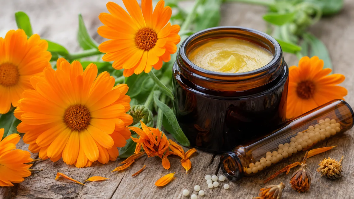 Ringelblumen-Globuli und eine geöffnete Tube Calendula-Salbe neben frischen orangefarbenen Ringelblüten auf hellem Holz