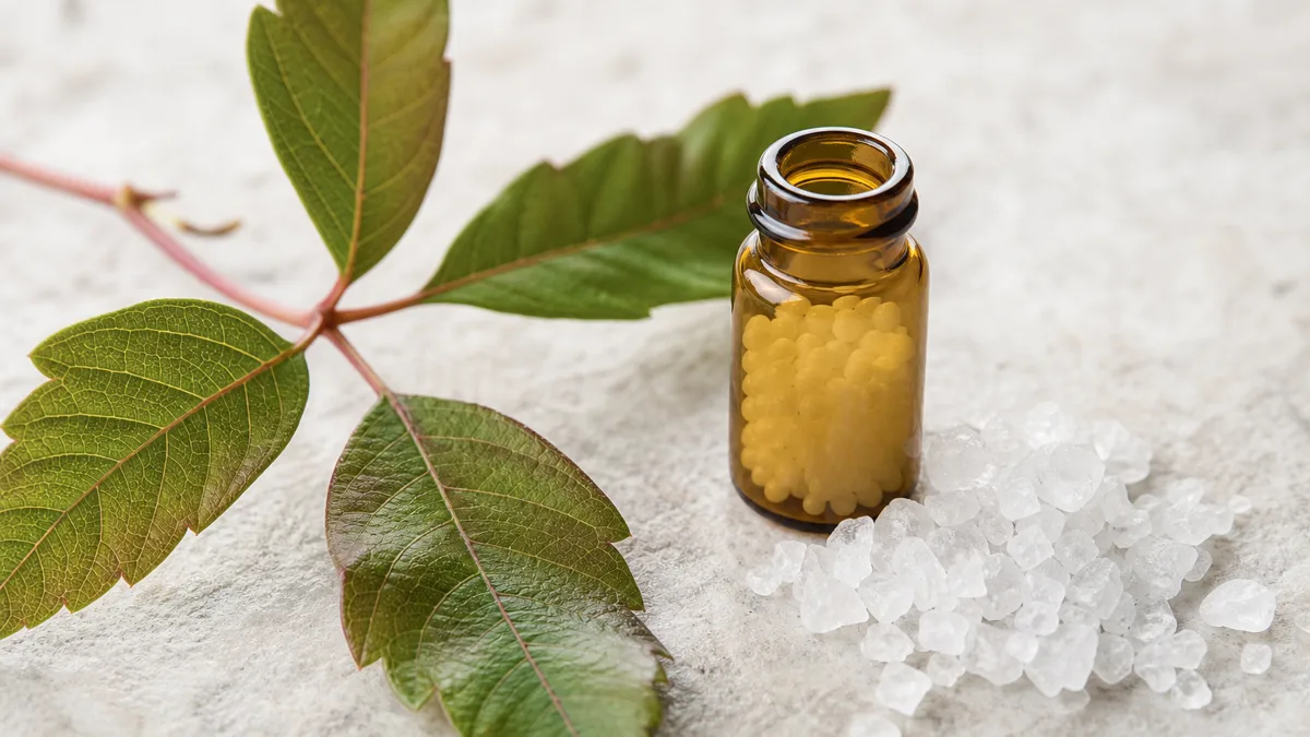 Röhrchen mit Globuli neben frischen Blättern von Rhus toxicodendron auf hellem Holz, im Hintergrund unscharf eine Person, die sich die kribbelnde Oberlippe berührt